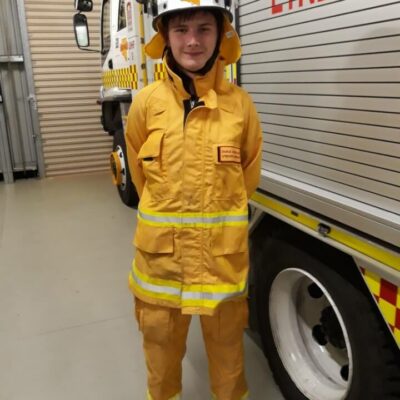 A young man in a yellow firefighters uniform and helmet stands beside a fire truck